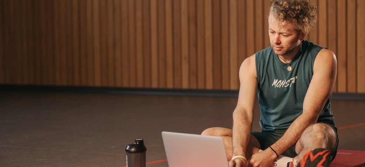 Man sitting on exercise mat with laptop, surrounded by dumbbells, creates a unique fitness workspace.